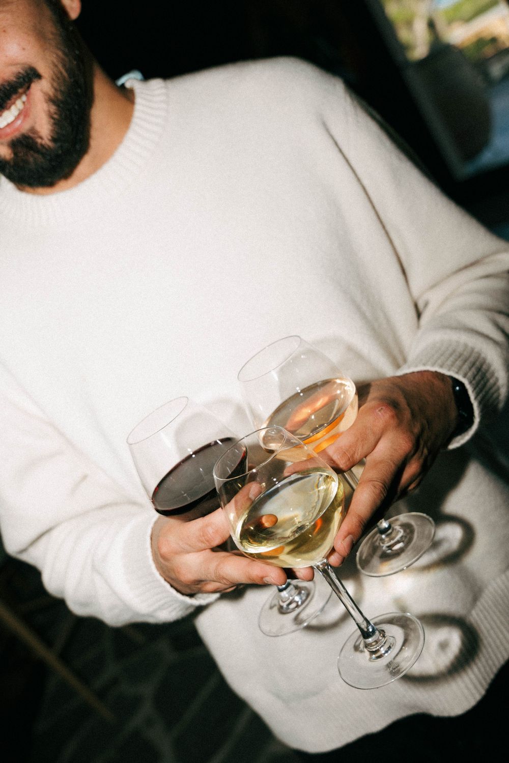 Smiling bearded man in white sweater holding multiple wine glasses with white and red wine at a social gathering.