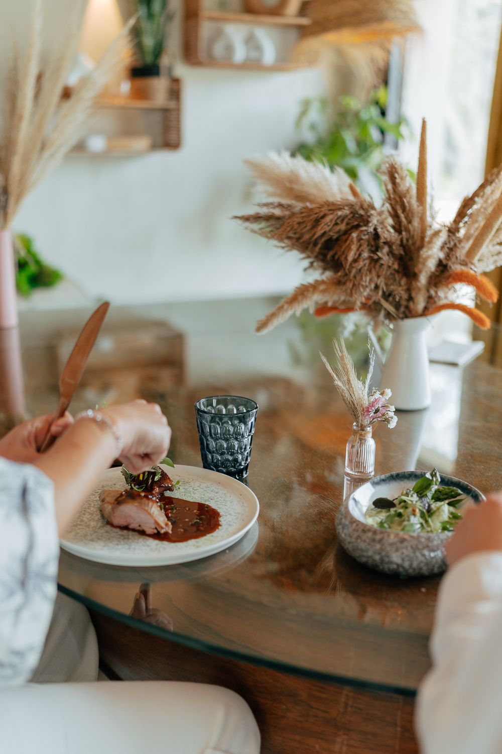 Person dining at a rustic wooden table with a gourmet meat dish, dried pampas grass vase, and small salad bowl.