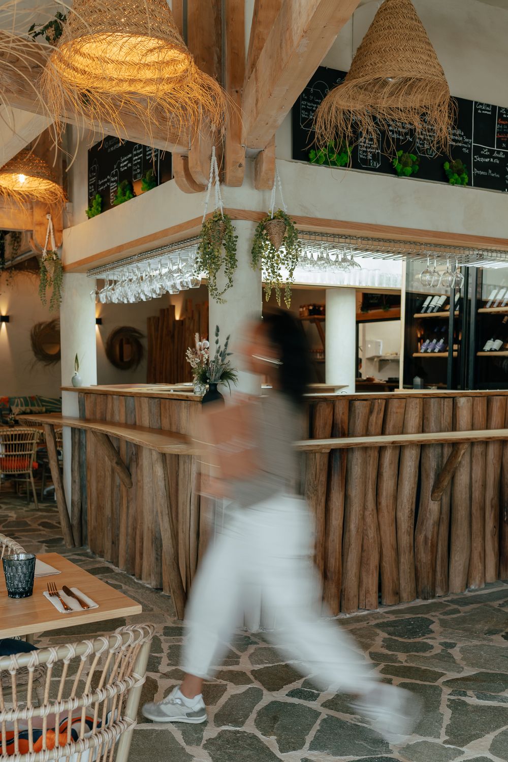 Blurred motion of a person walking through a rustic restaurant with wooden bar, wicker lamps, and stone floor.