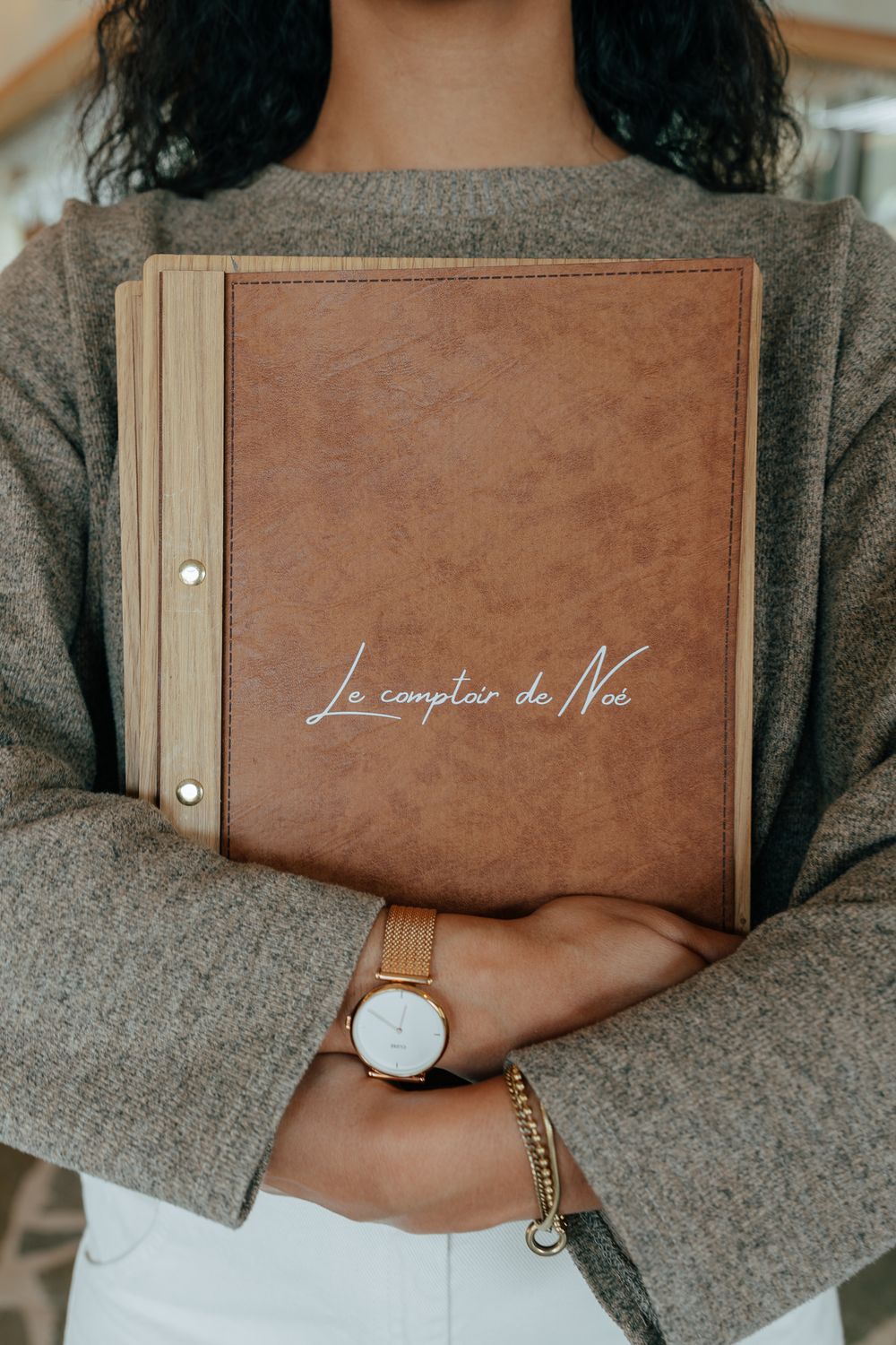 Woman holding a leather-bound menu for Le Comptoir de Noé, wearing a gold watch and gray sweater.