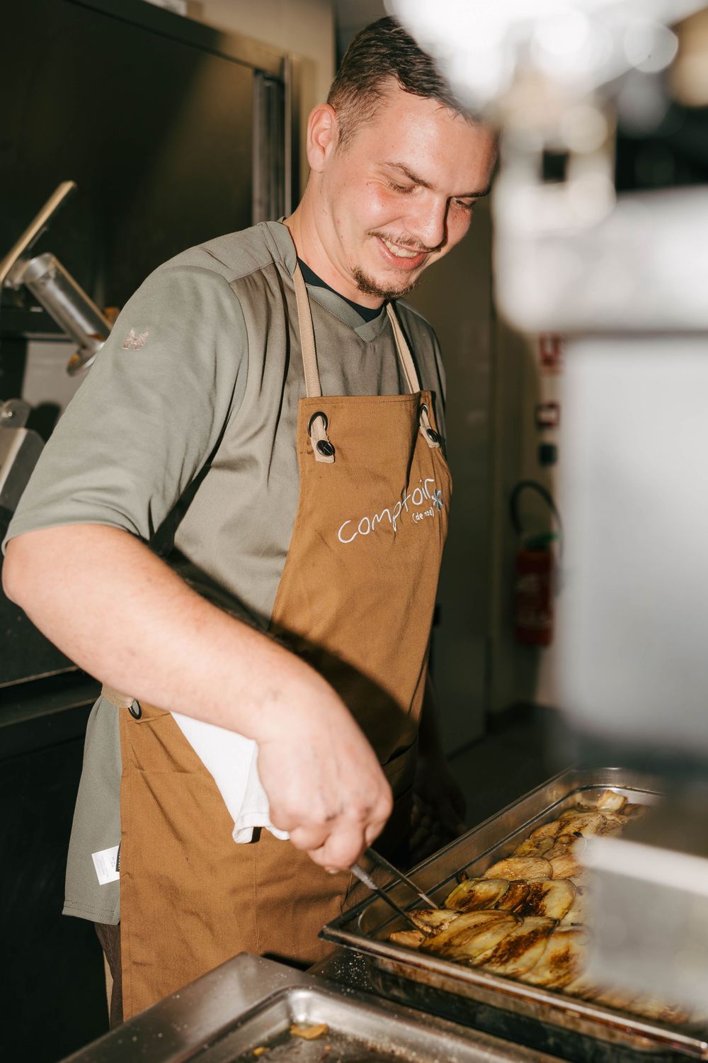 Smiling chef in brown apron uses tongs to cook golden potatoes in a professional kitchen.