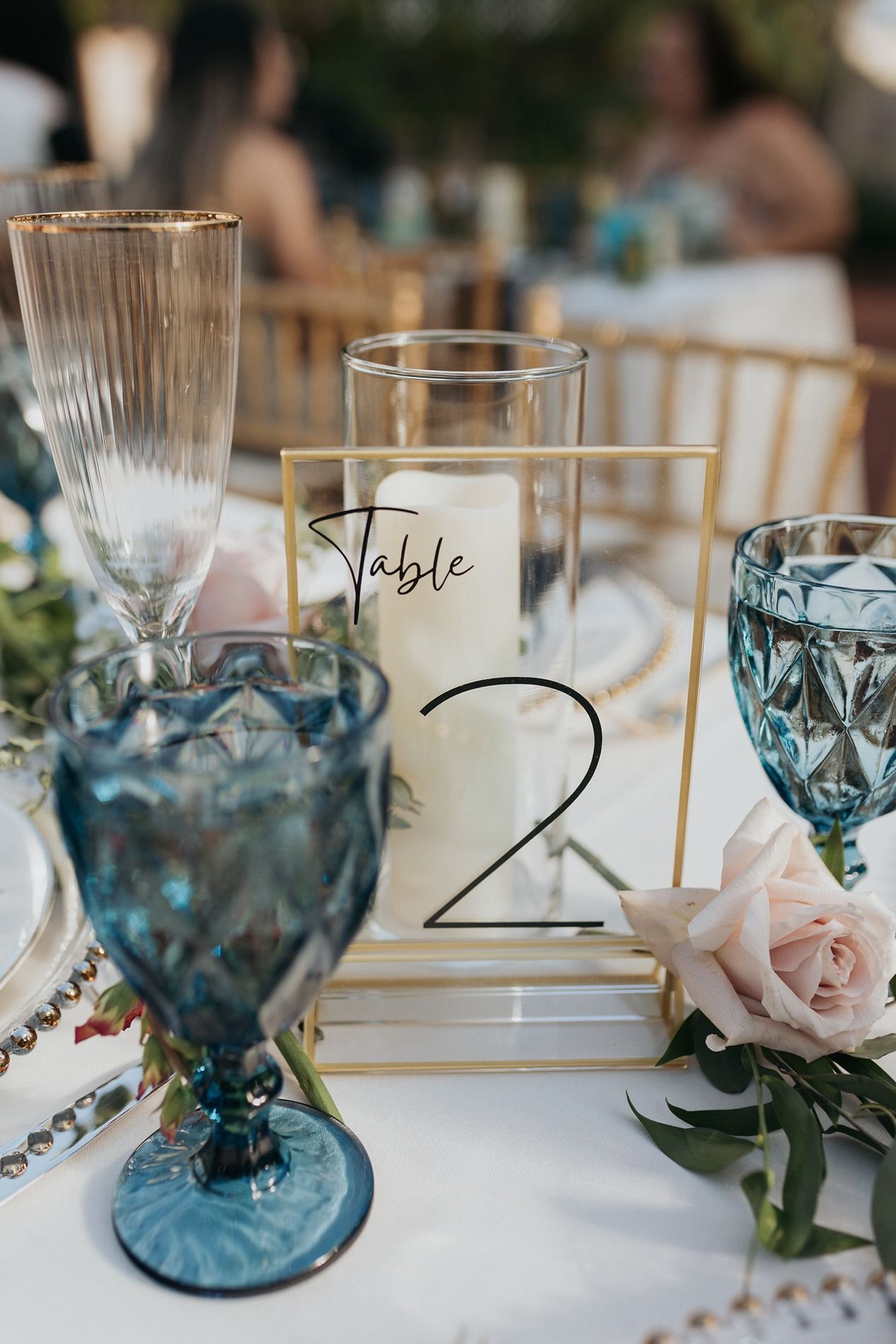 Elegant wedding table setting with blue glassware, gold accents, candle, pink rose, and Table 2 sign.