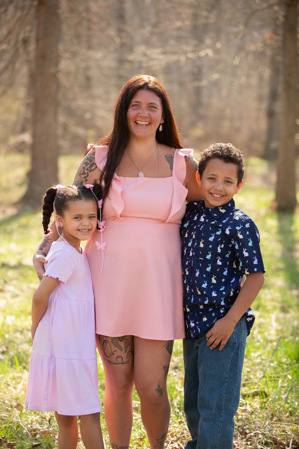 A smiling woman in a pink dress poses outdoors with two young children in a sunlit forest setting.