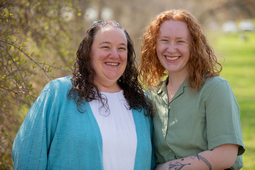 Two smiling women with curly hair pose together outdoors in a green park setting on a sunny day.