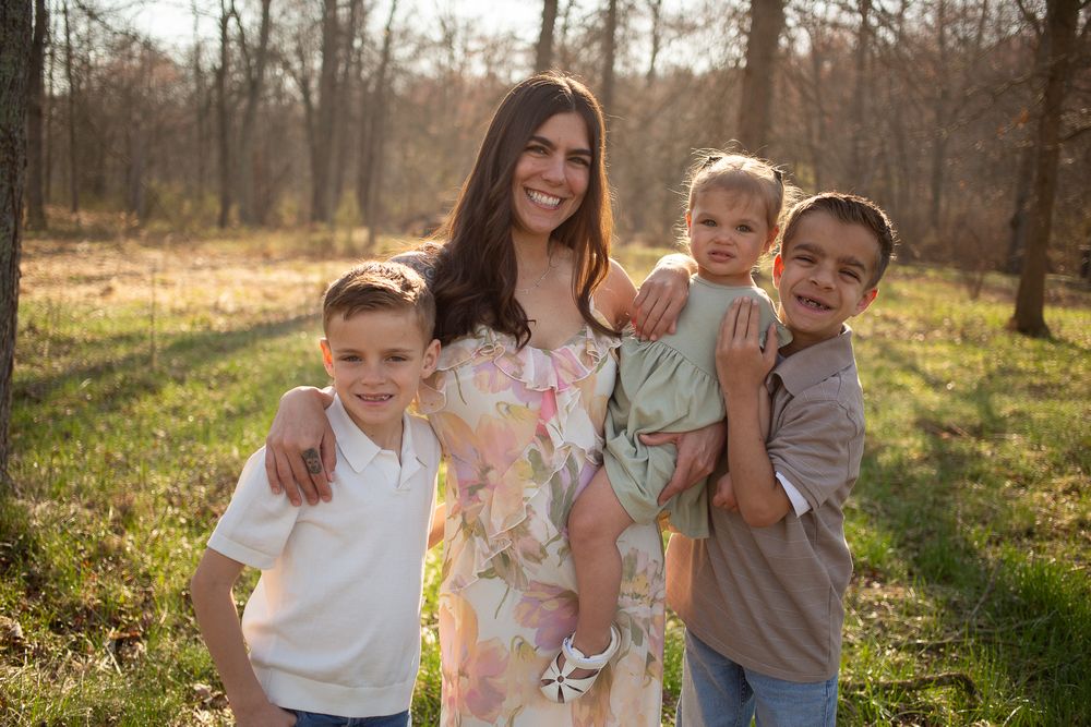 A smiling woman in a floral dress holds a toddler while two boys pose beside her outdoors in a sunny wooded setting.