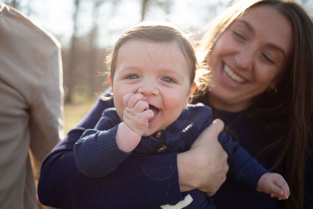 Smiling baby in navy outfit held by laughing woman outdoors in warm sunlight.