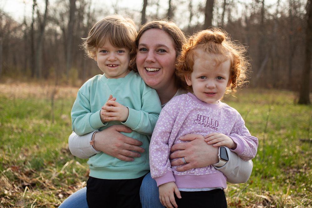 A smiling mother hugs her two young toddler girls outdoors in a field during fall.