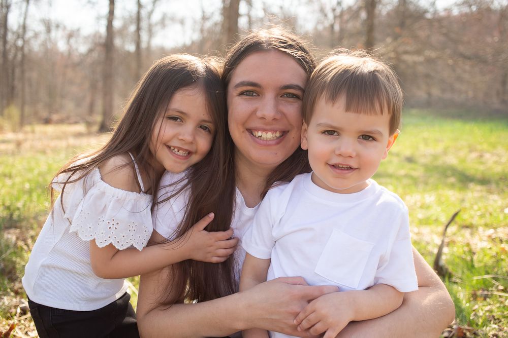 A smiling mother poses with her young daughter and son in white outfits in a sunny park.