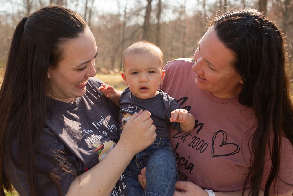 Two women smiling and holding a baby outdoors in a sunny park setting during a family photo session.