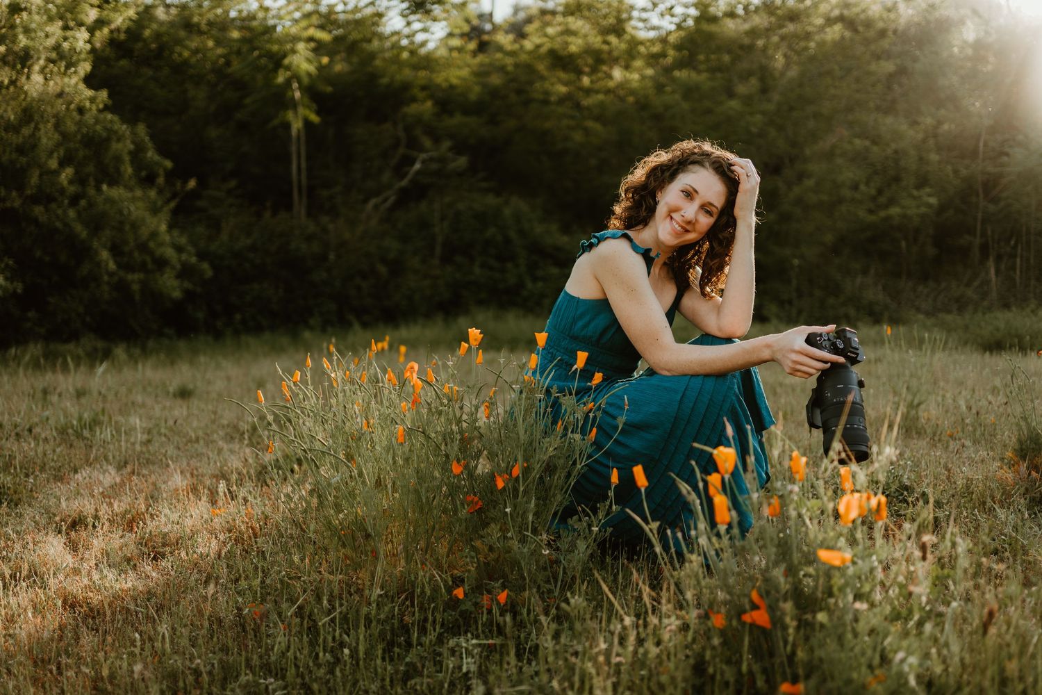 Woman in teal dress sitting in a sunlit meadow holding a camera, surrounded by wildflowers and golden light.