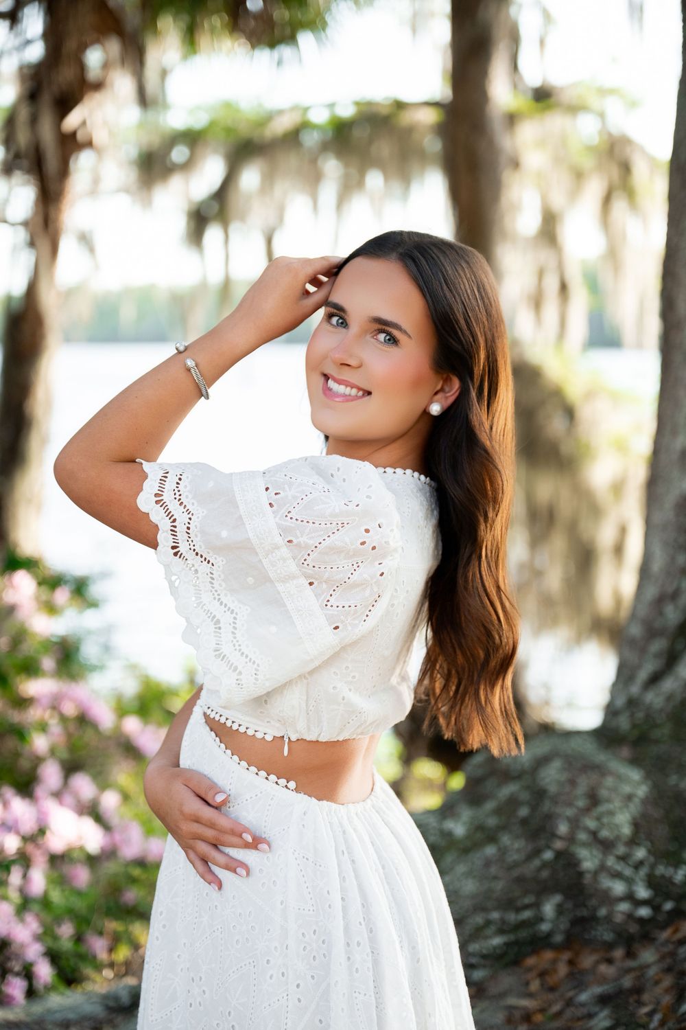 Young woman in white eyelet crop top and skirt posing outdoors near trees and flowers.