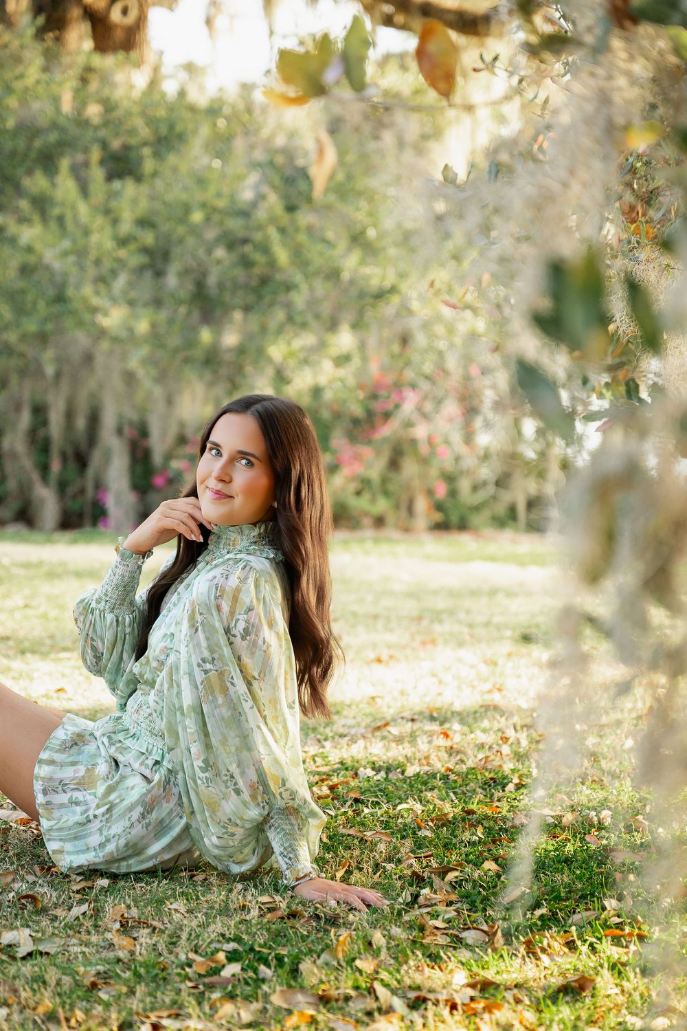 Young woman in floral dress sitting on grass under a tree in a lush green park, looking up dreamily.