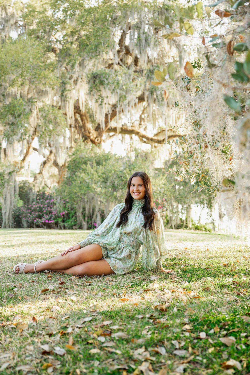 Smiling young woman in green floral mini dress sitting on grass beneath moss-draped oak trees outdoors.