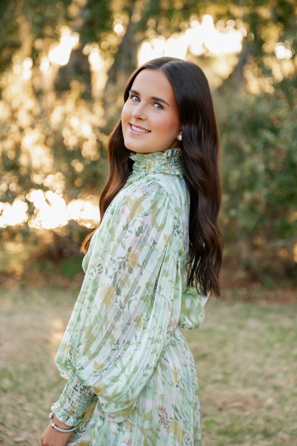 Young woman in green floral dress smiling over shoulder outdoors with sunlit trees in background.