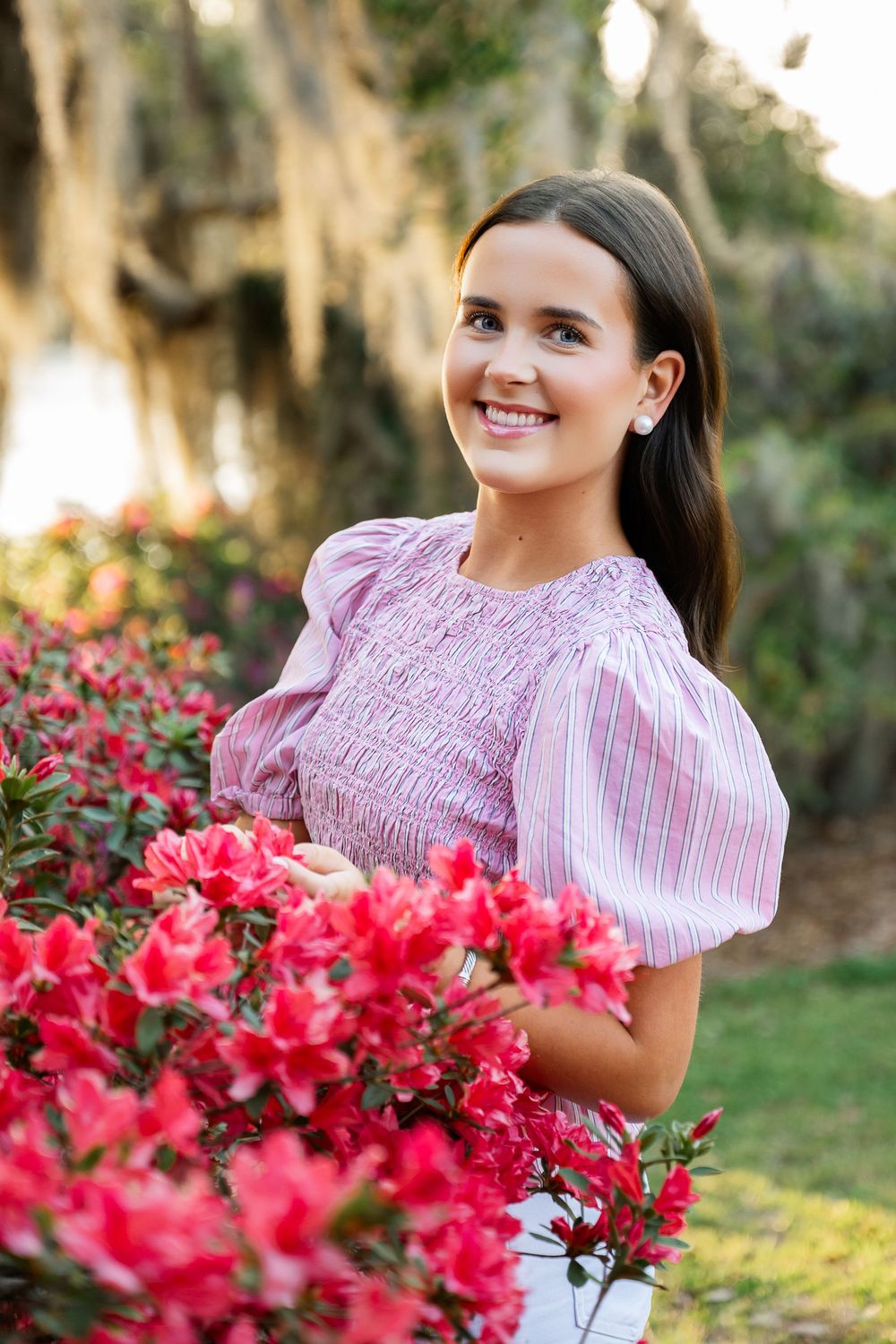 Young woman in pink striped smocked top smiling among vibrant red azalea blooms in a lush garden.