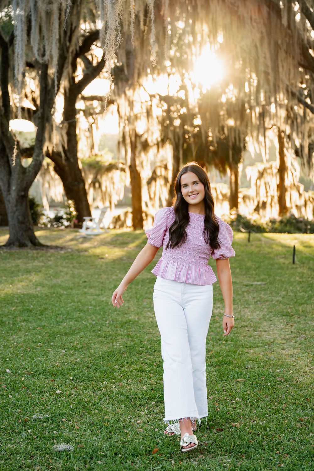 Smiling brunette woman in pink peplum top and white pants walking in park with Spanish moss trees at sunset.