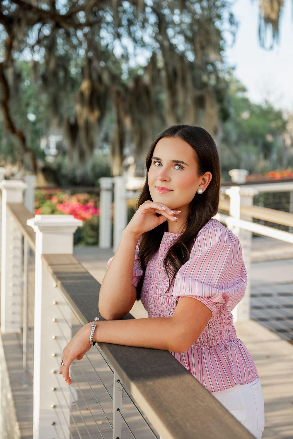Young brunette woman in pink striped smocked top leaning on a white railing on a sunny outdoor deck.