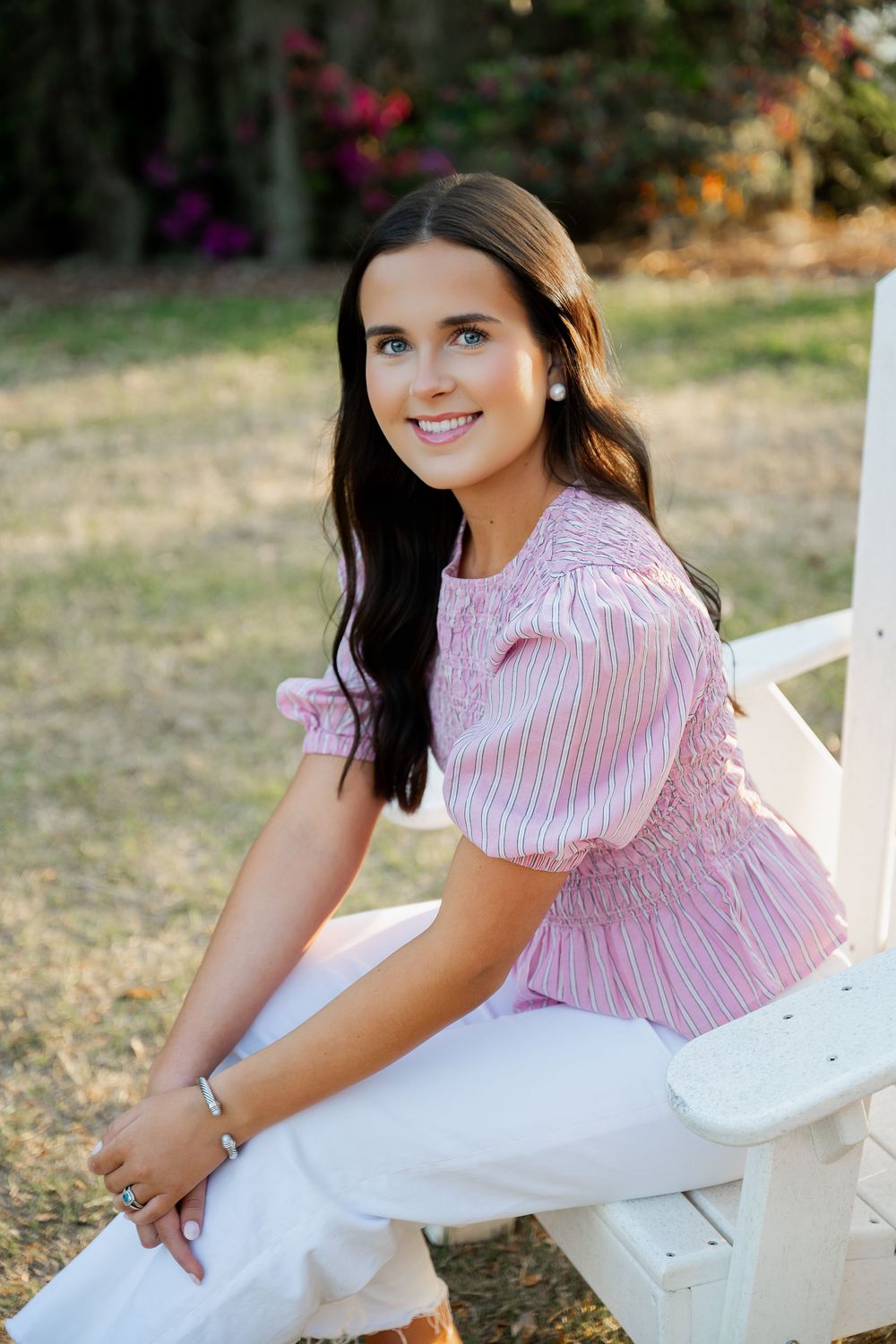 Smiling young woman in pink striped smocked blouse and white pants sitting on white bench in garden.