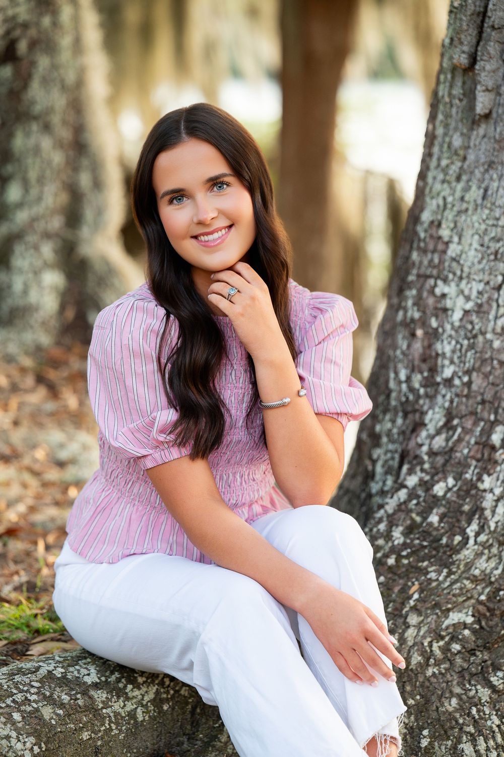 Young woman in pink striped top and white pants sitting by a tree, smiling outdoors.