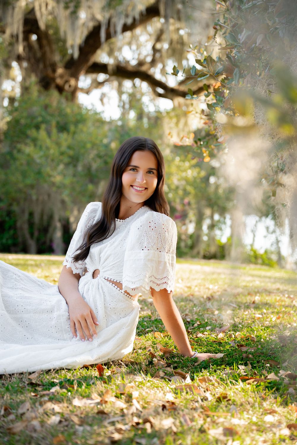 Young woman in white eyelet dress sitting gracefully on grass beneath moss-draped oak trees in sunny park.