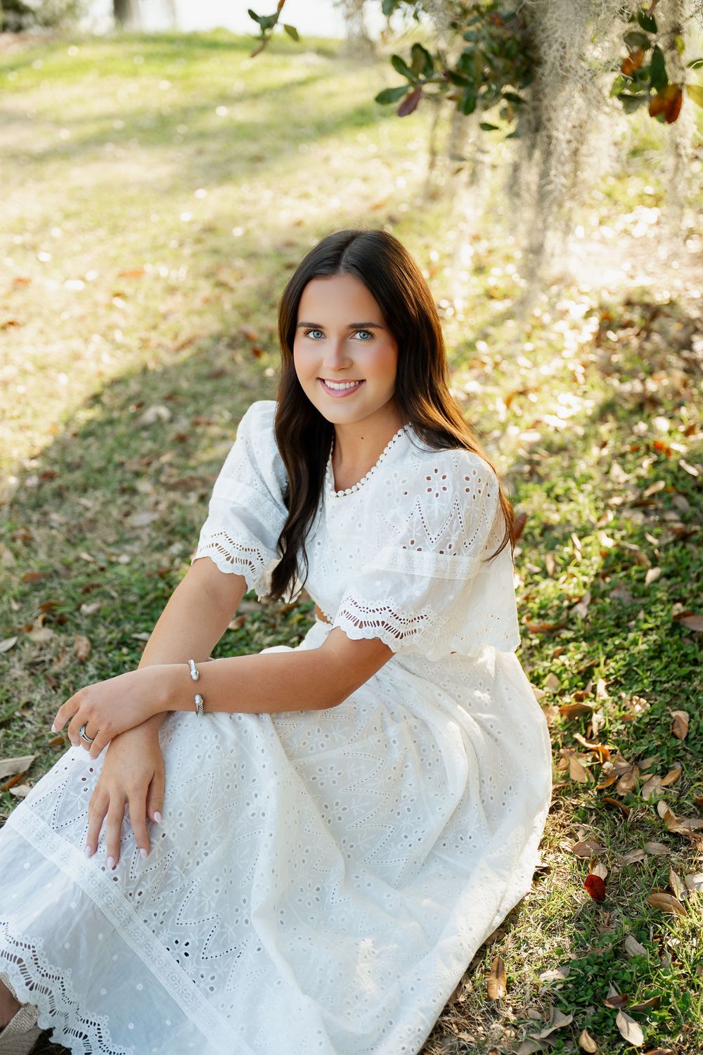 Young woman in white eyelet dress sitting outdoors under a tree, smiling warmly in a sunlit garden setting.