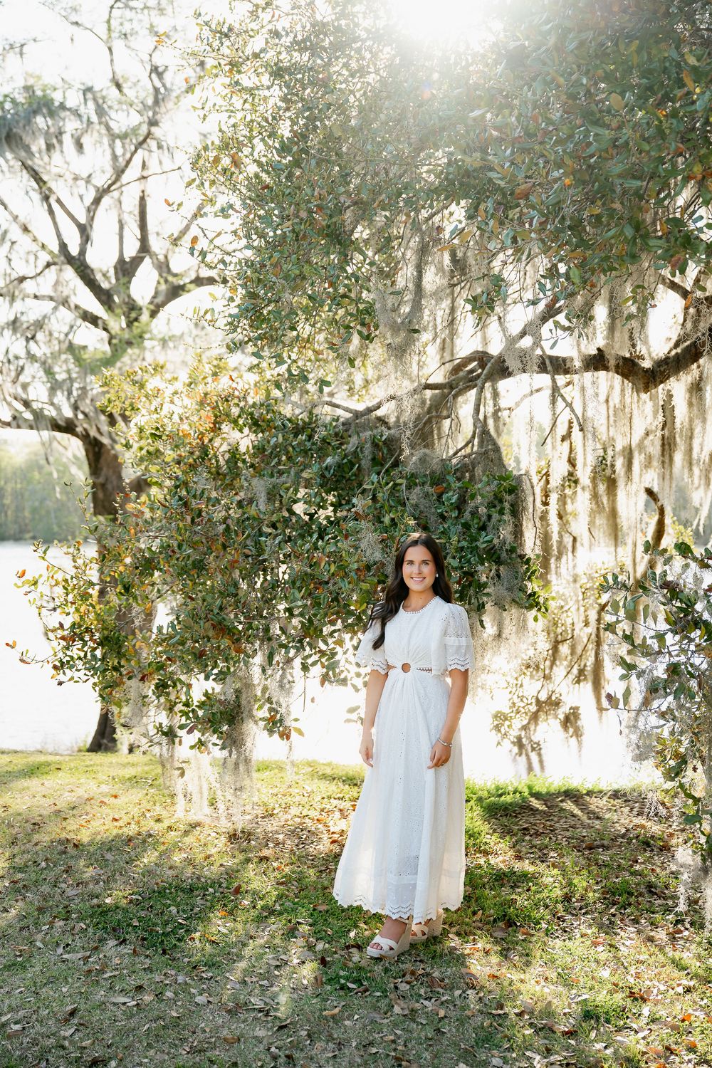 Smiling young woman in white midi dress standing under Spanish moss-covered oak tree near sunlit lake.
