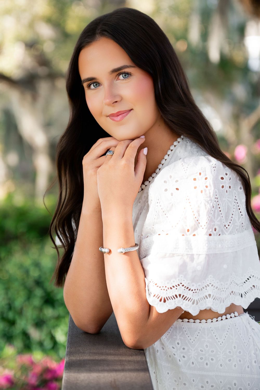 Young woman in white eyelet dress poses outdoors, wearing pearl bracelet and ring, with lush green trees behind her.