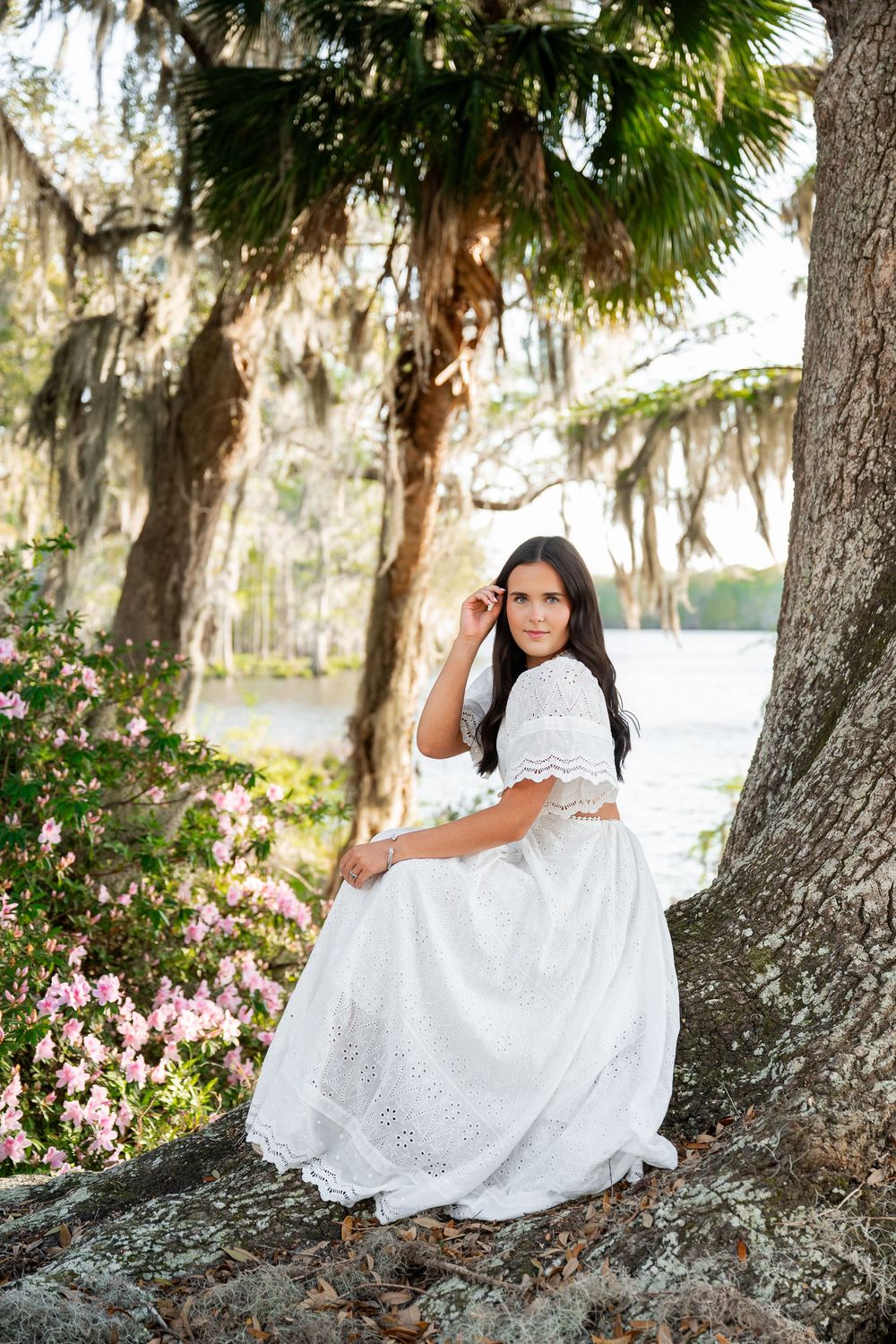 Elegant young woman in white lace dress posing by a large tree with Spanish moss and pink azaleas nearby.
