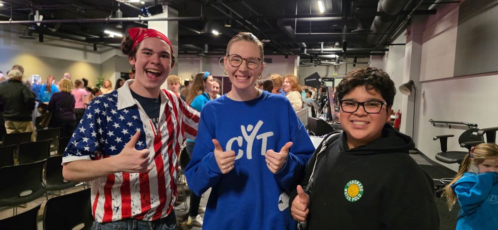 Three smiling teens giving thumbs up at a youth group event in a large indoor venue.