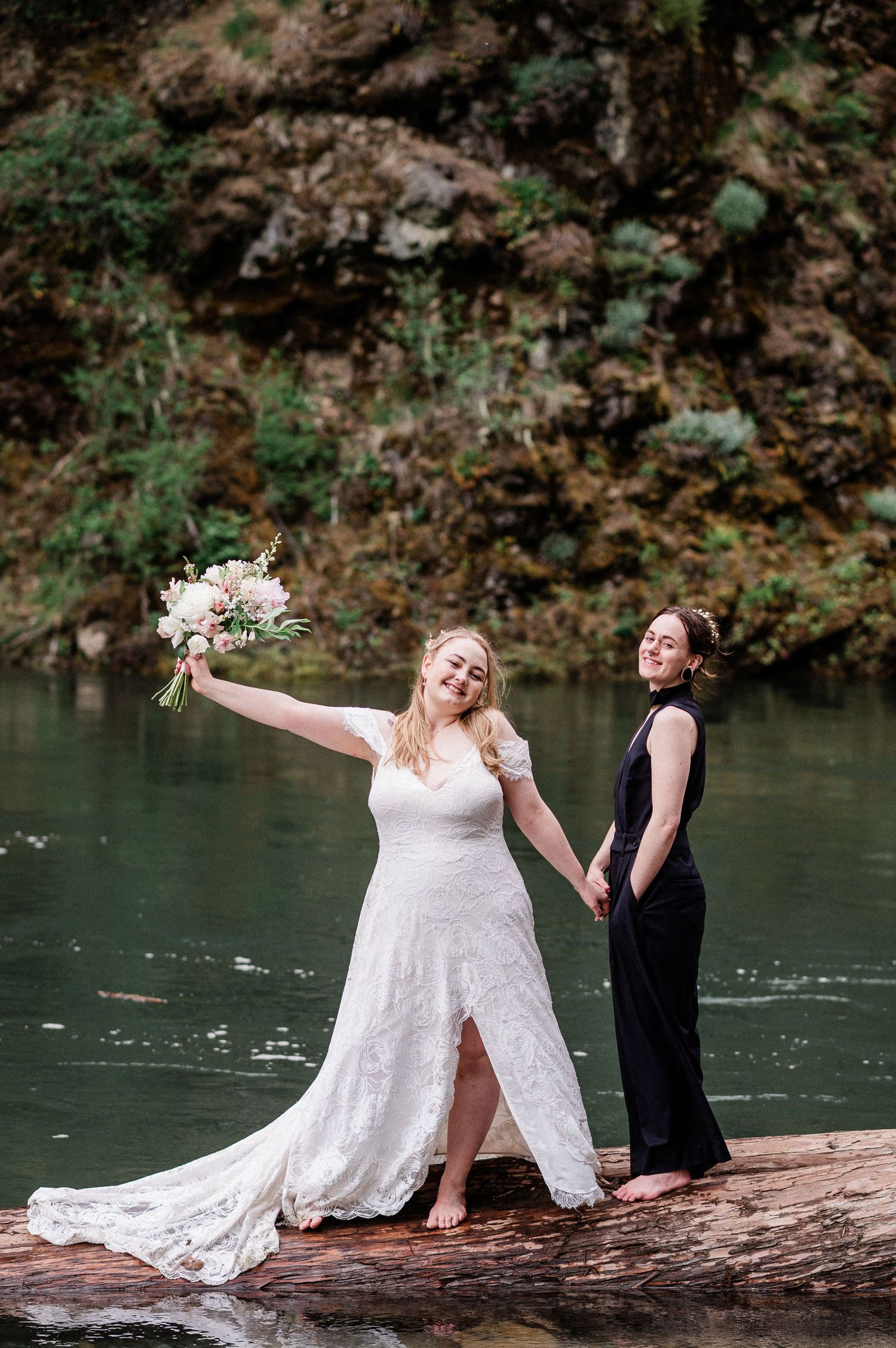 Two brides on a fallen log by a serene river, one wearing an elegant white lace gown with a flowing train and the other a black pantsuit
