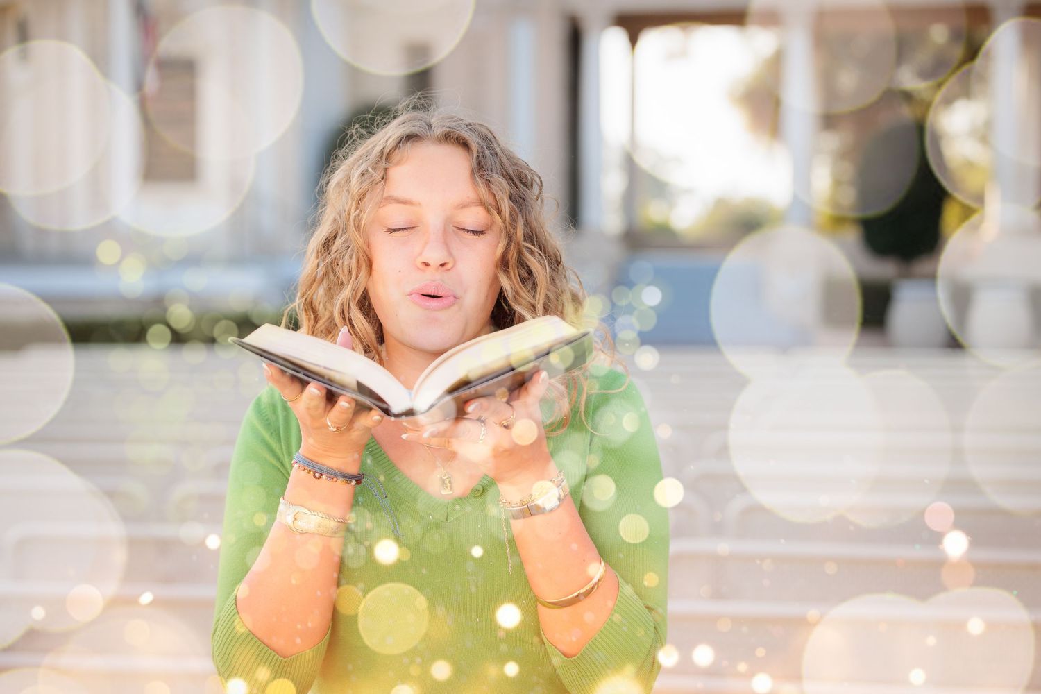 Woman in green sweater blowing pages of an open book with sparkling bokeh light effects around her.