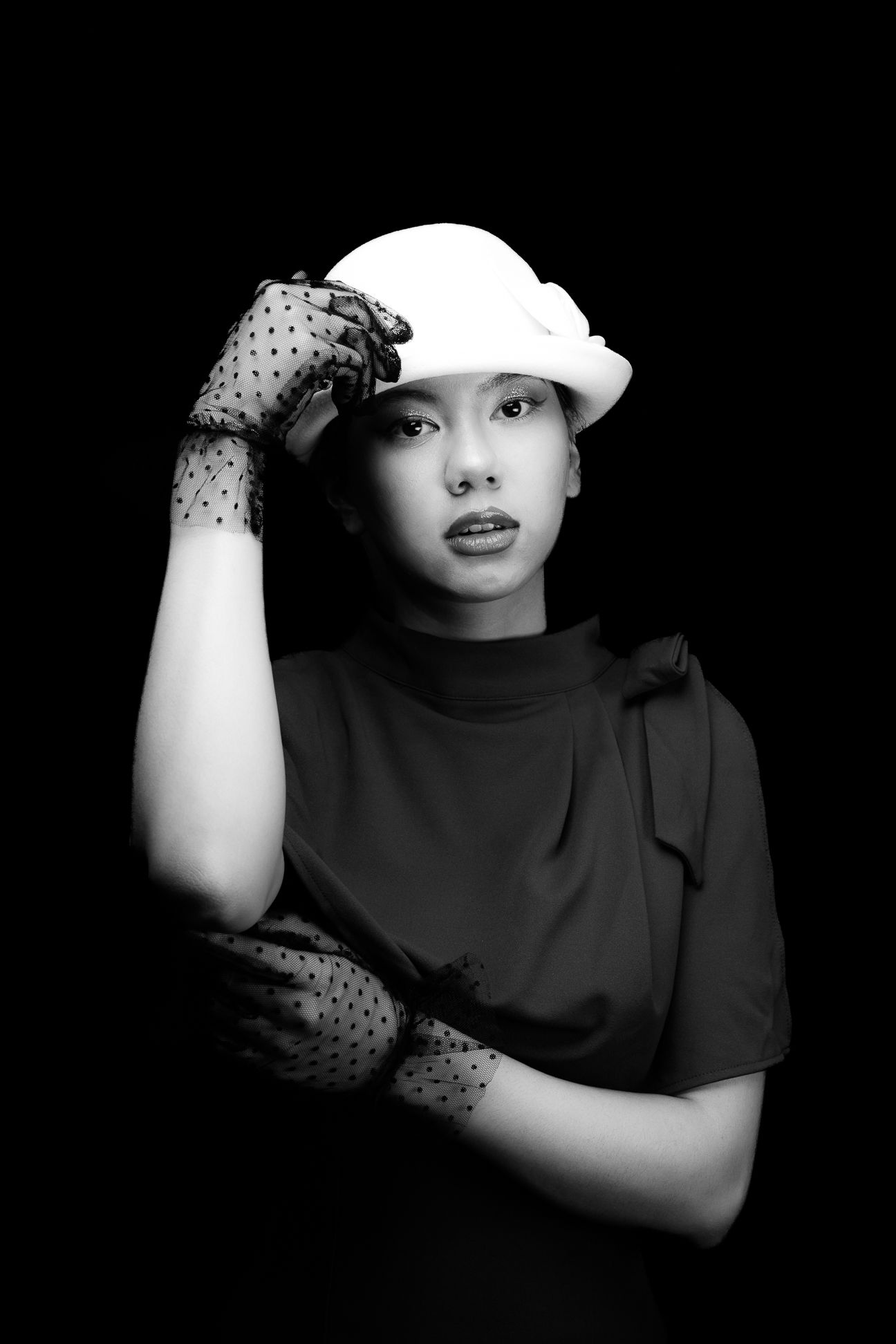 Black and white image of a girl in a white, wool, bowler hat with polka dot, clear black gloves, holding her arm and hat, looking straight