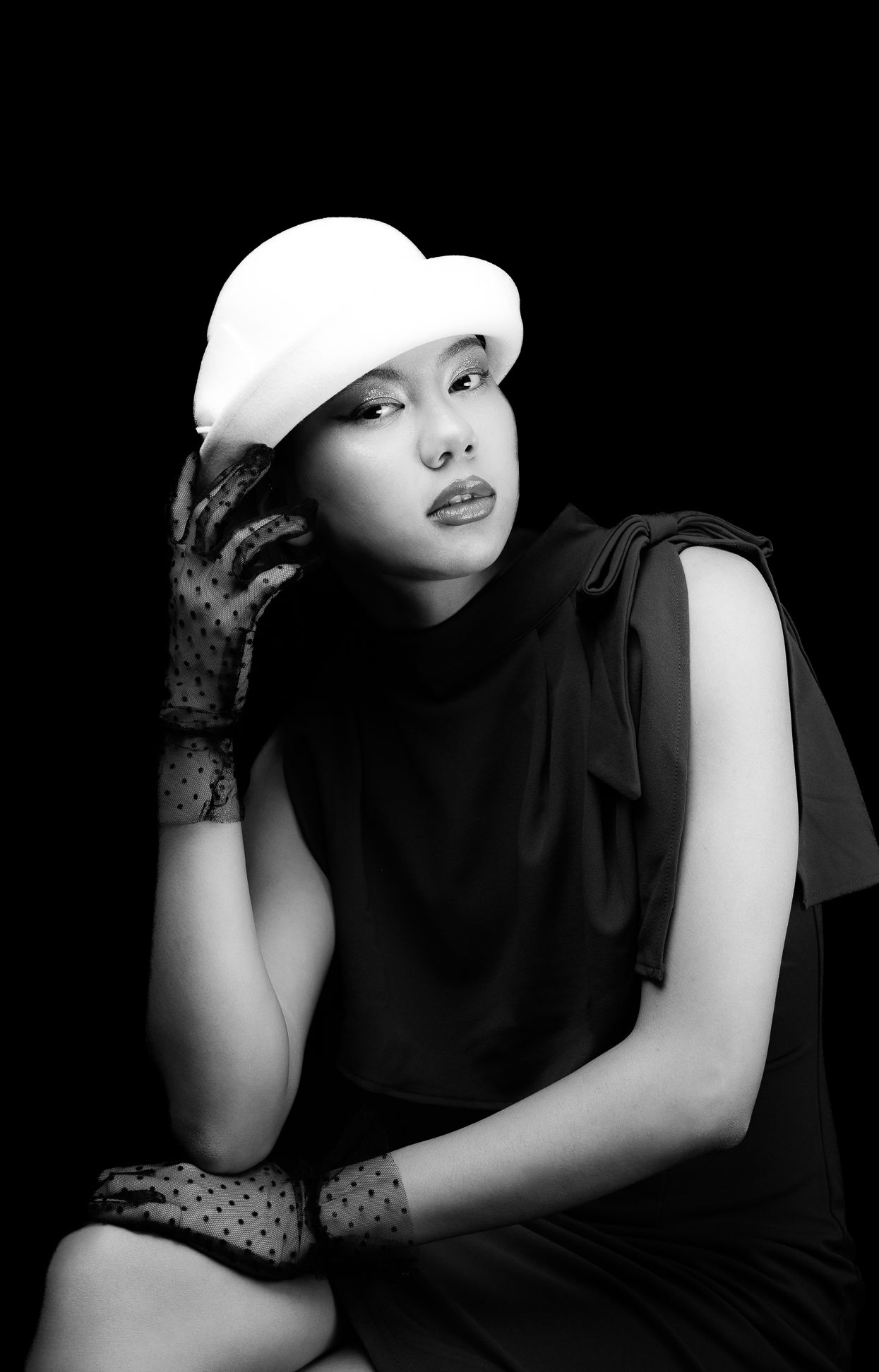 Black and white image of a girl in a white, wool, bowelr hat with polka dot clear black gloves, leaning over and looking at the camera