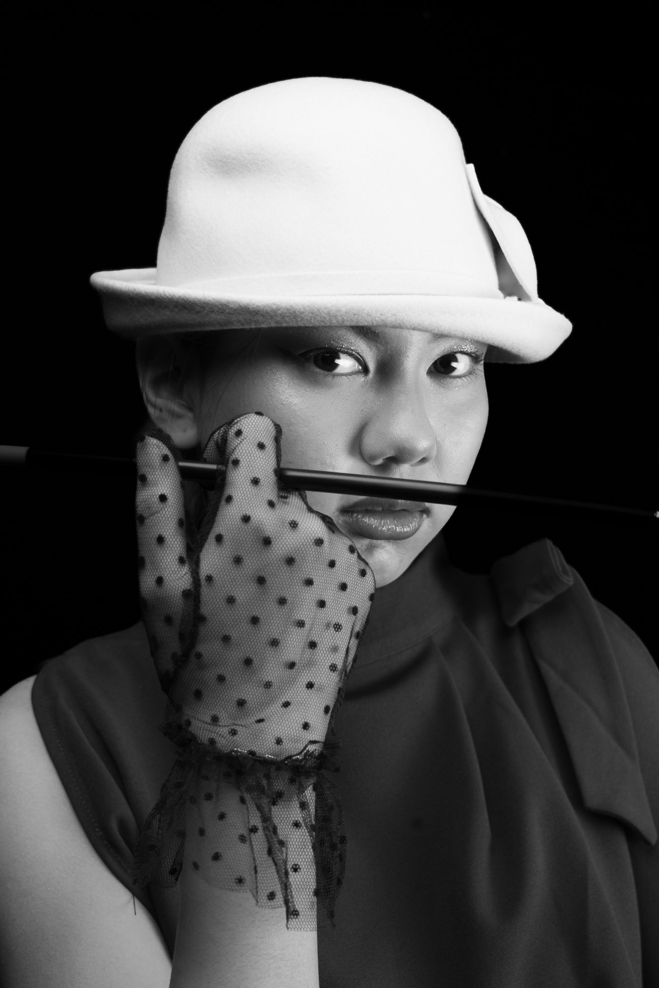 Black and white image of a girl in a white, wool, bowler hat with polka dot, clear black gloves, holding up a cigarette holder