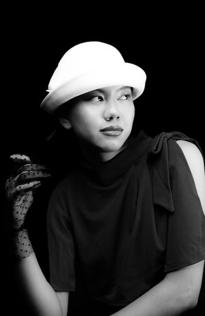 Black and white image of a girl in a white, wool, bowler hat with polka dot, clear black gloves, holding a cigarette, looking right
