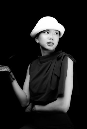 Black and white image of a girl in a white, wool, bowler hat with polka dot, clear black gloves, holding a cigarette on side, looking right