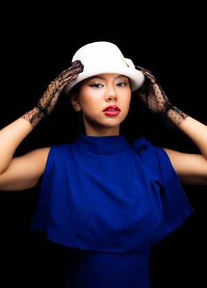 Image of a girl in a white, wool, bowelr hat with polka dot clear black gloves, leaning over and looking at the camera, with hands up on hat
