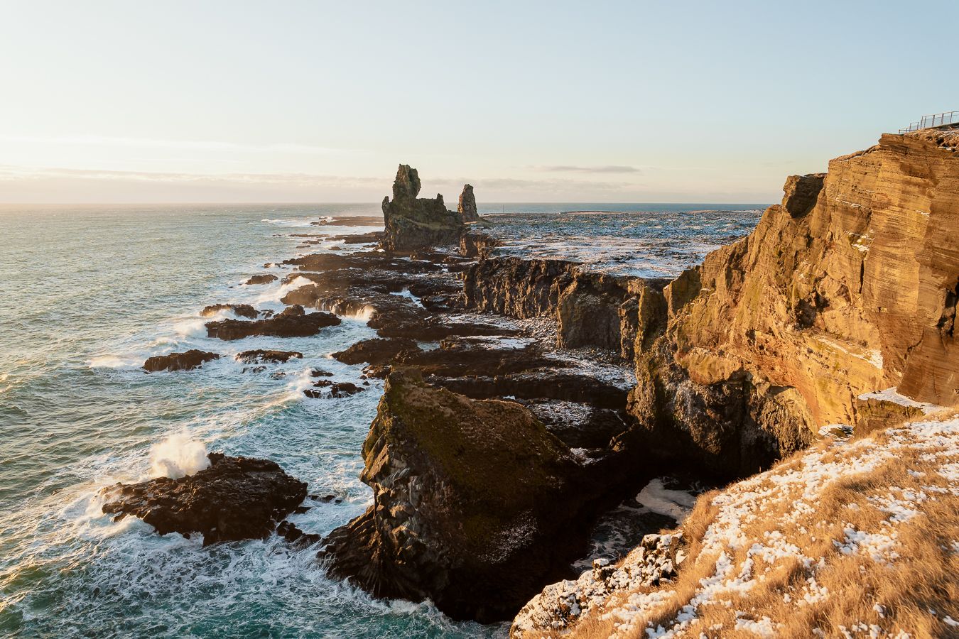 Rocky coastal cliffs with dramatic sea stacks rising from turbulent ocean waves at golden hour in Iceland.