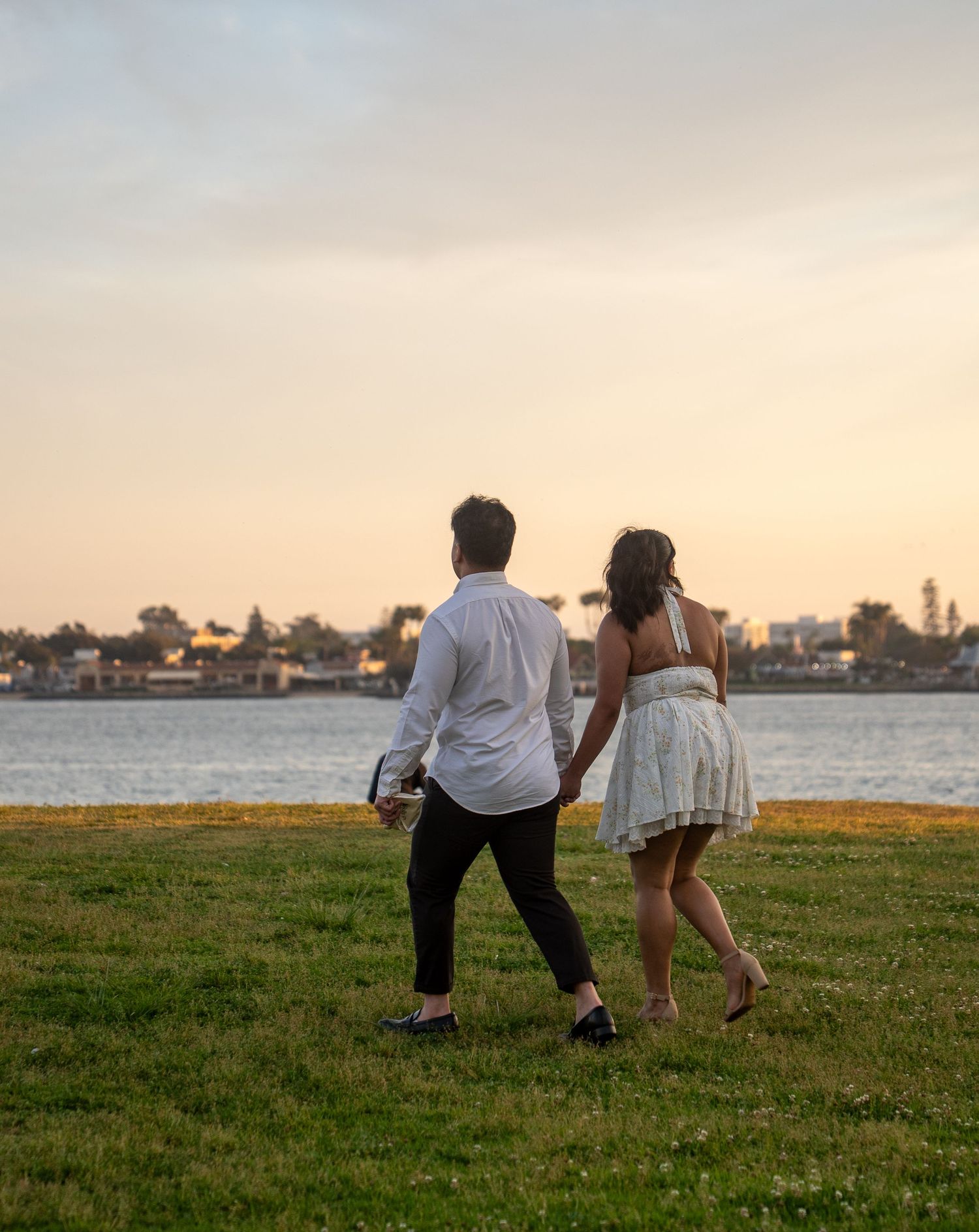 Couple holding hands walking on green grass toward a scenic waterfront at sunset, wearing white outfits.