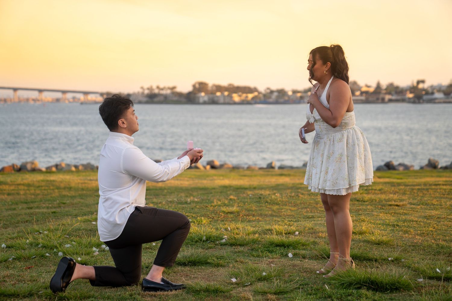 Man proposing on one knee to woman at sunset by the waterfront, romantic marriage proposal outdoors.