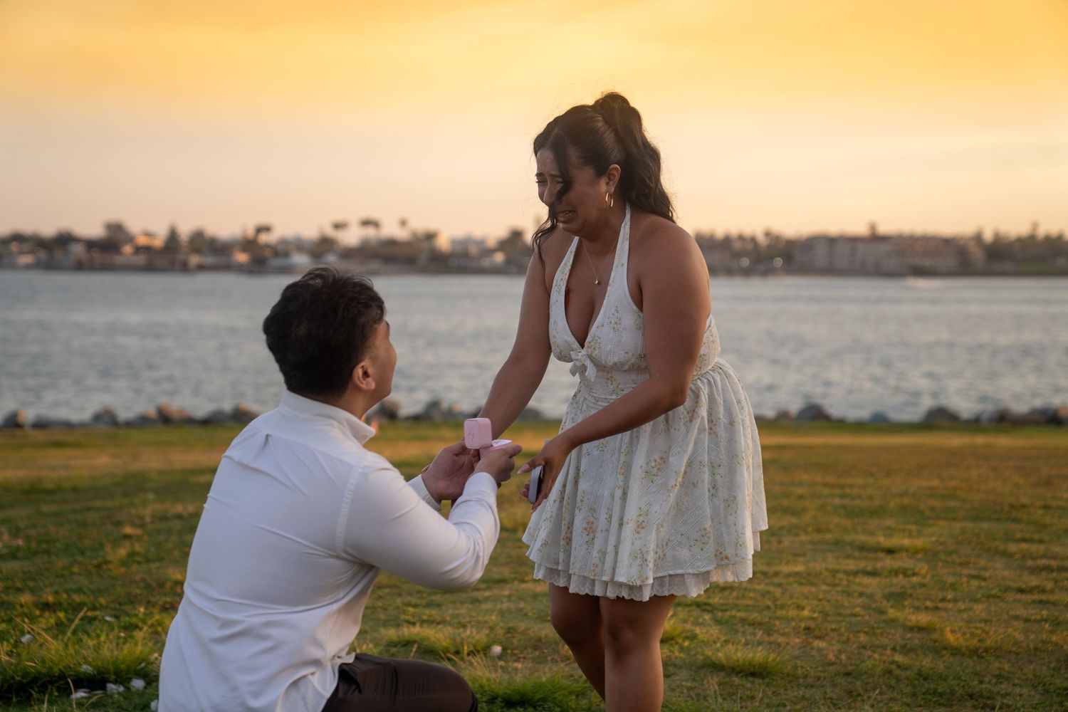 Man kneeling to propose to woman at sunset by the water, romantic marriage proposal on green grass.