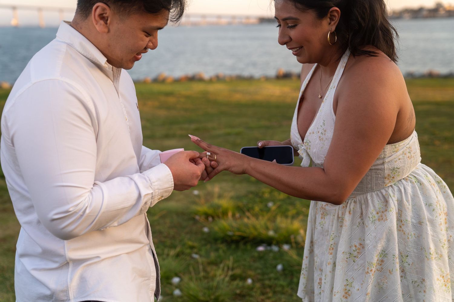 Couple exchanging rings outdoors near waterfront at sunset during romantic marriage proposal ceremony.