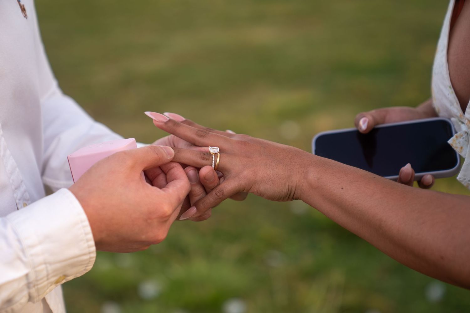 Close-up of a couple exchanging wedding rings outdoors, with one partner sliding a gold ring onto the other's finger.