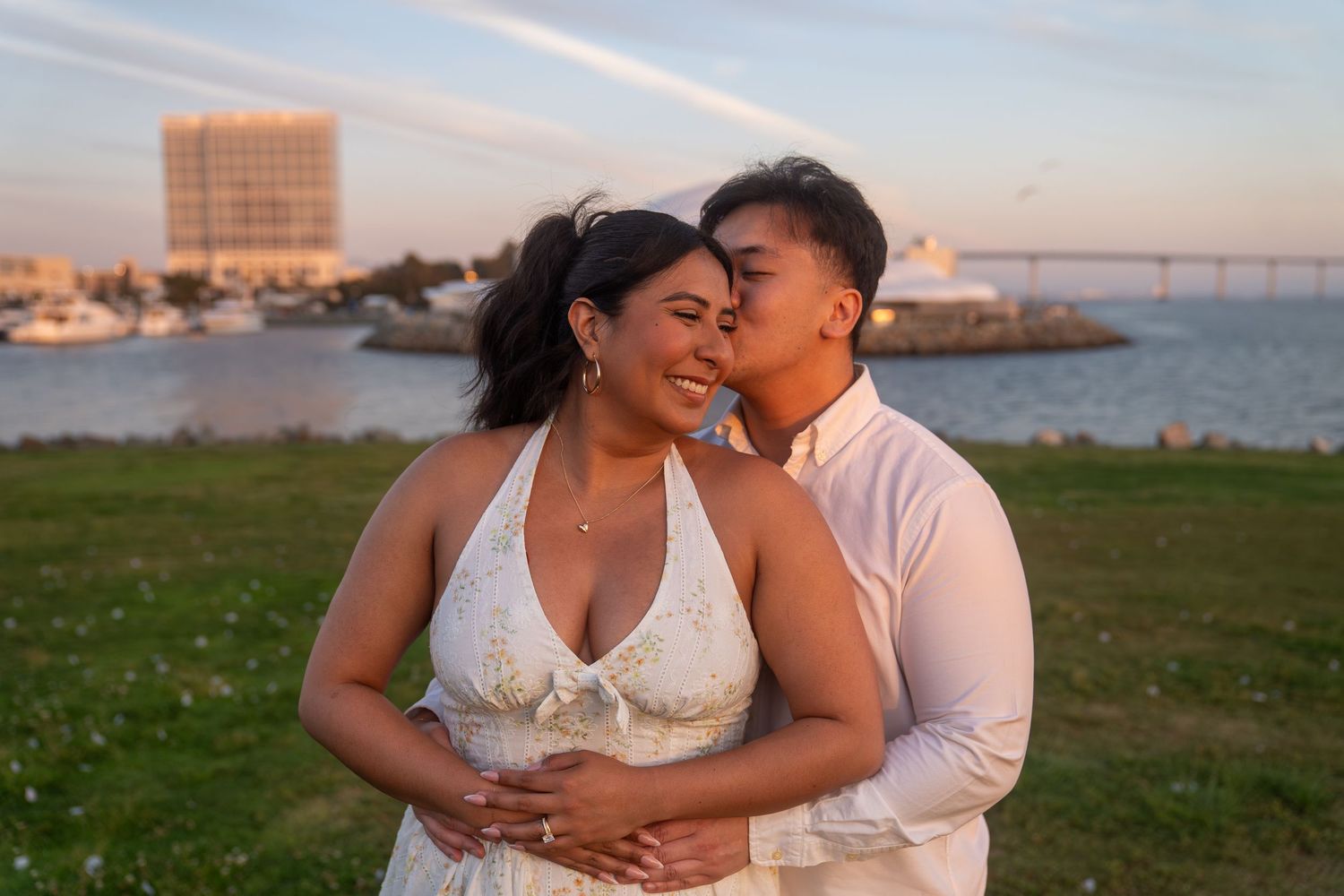 Couple embracing at sunset near a waterfront, woman in white halter wedding dress, man in light pink shirt, marina in background.