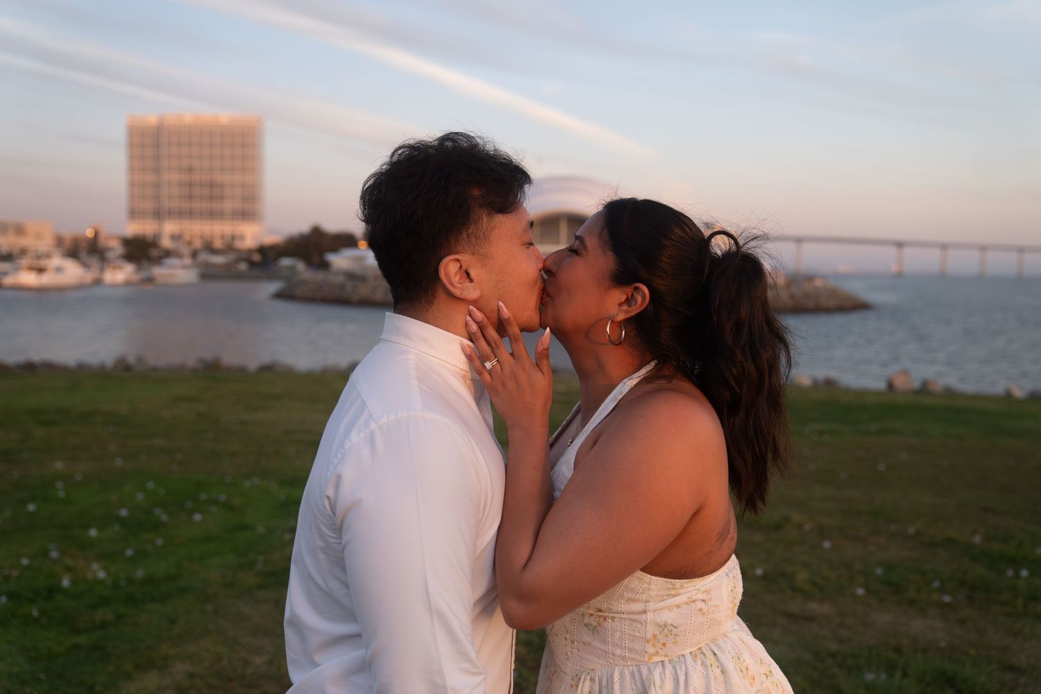 Couple sharing a romantic kiss at sunset near the waterfront with a bridge visible in the background.