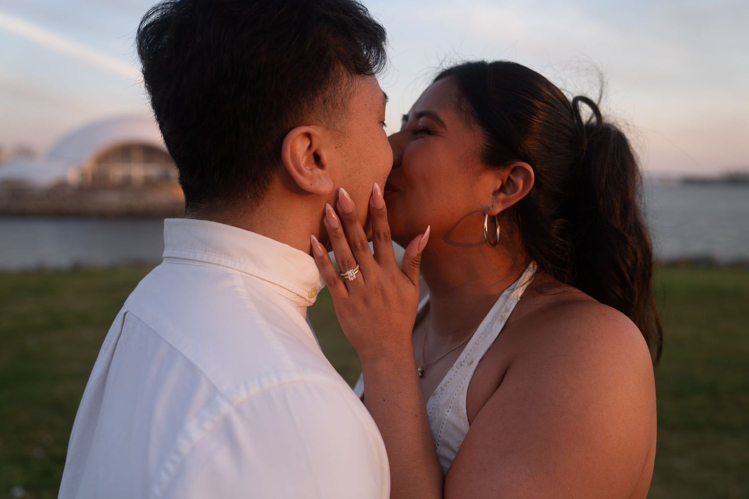 Couple sharing a romantic kiss at sunset near the waterfront, woman wearing a white dress and engagement ring.