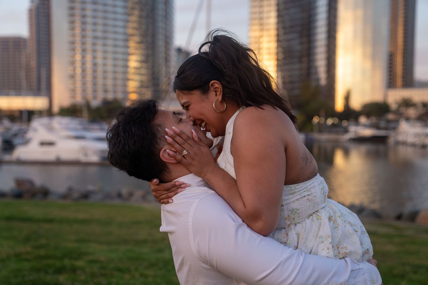 Couple sharing a romantic kiss at sunset near a marina with city skyline in the background.