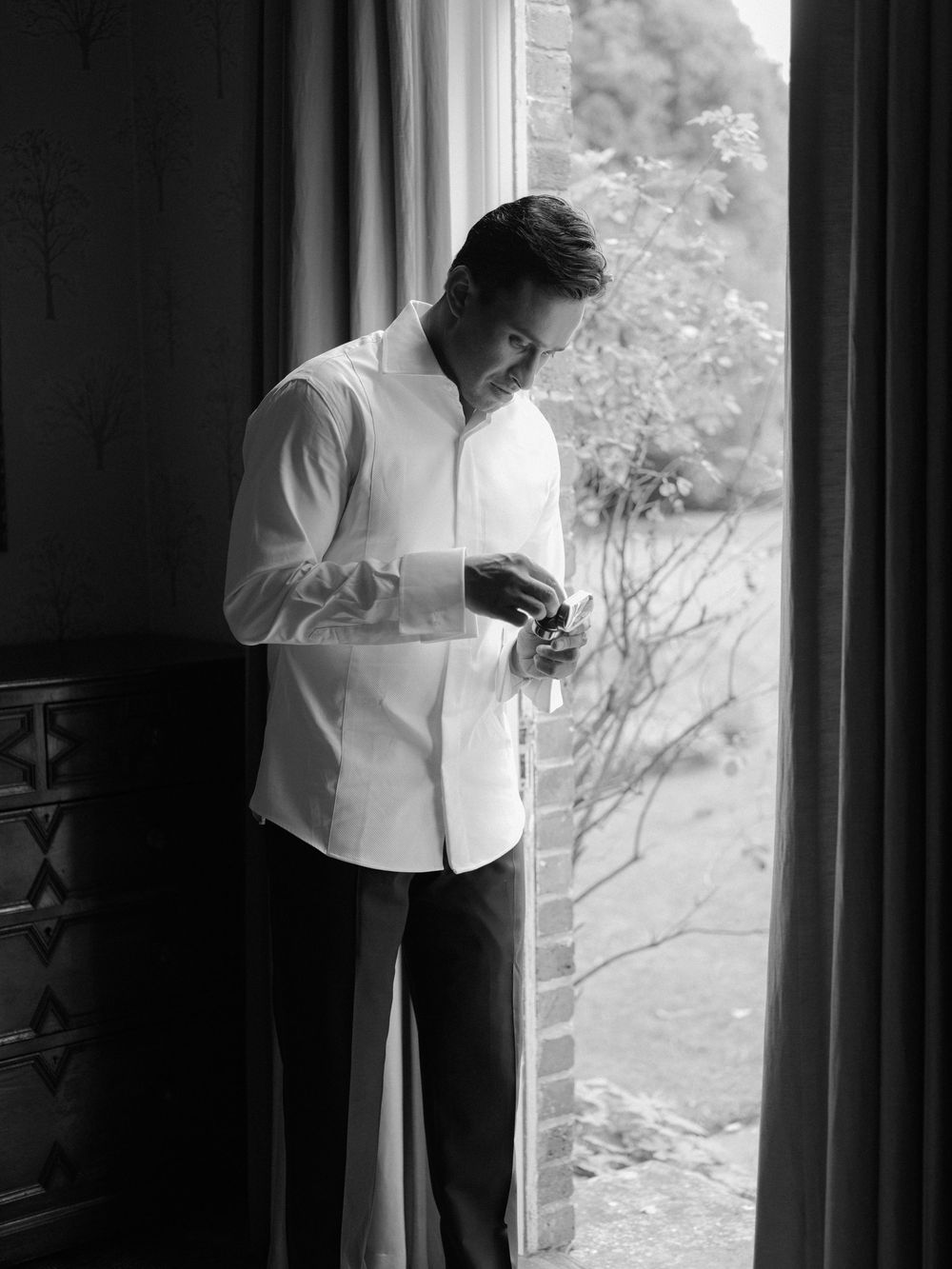 Groom adjusting cufflinks by window in black and white wedding preparation photo.