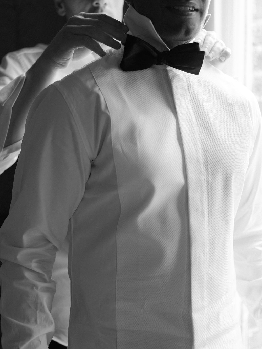 Close-up black and white photo of groom having bow tie adjusted before wedding ceremony.