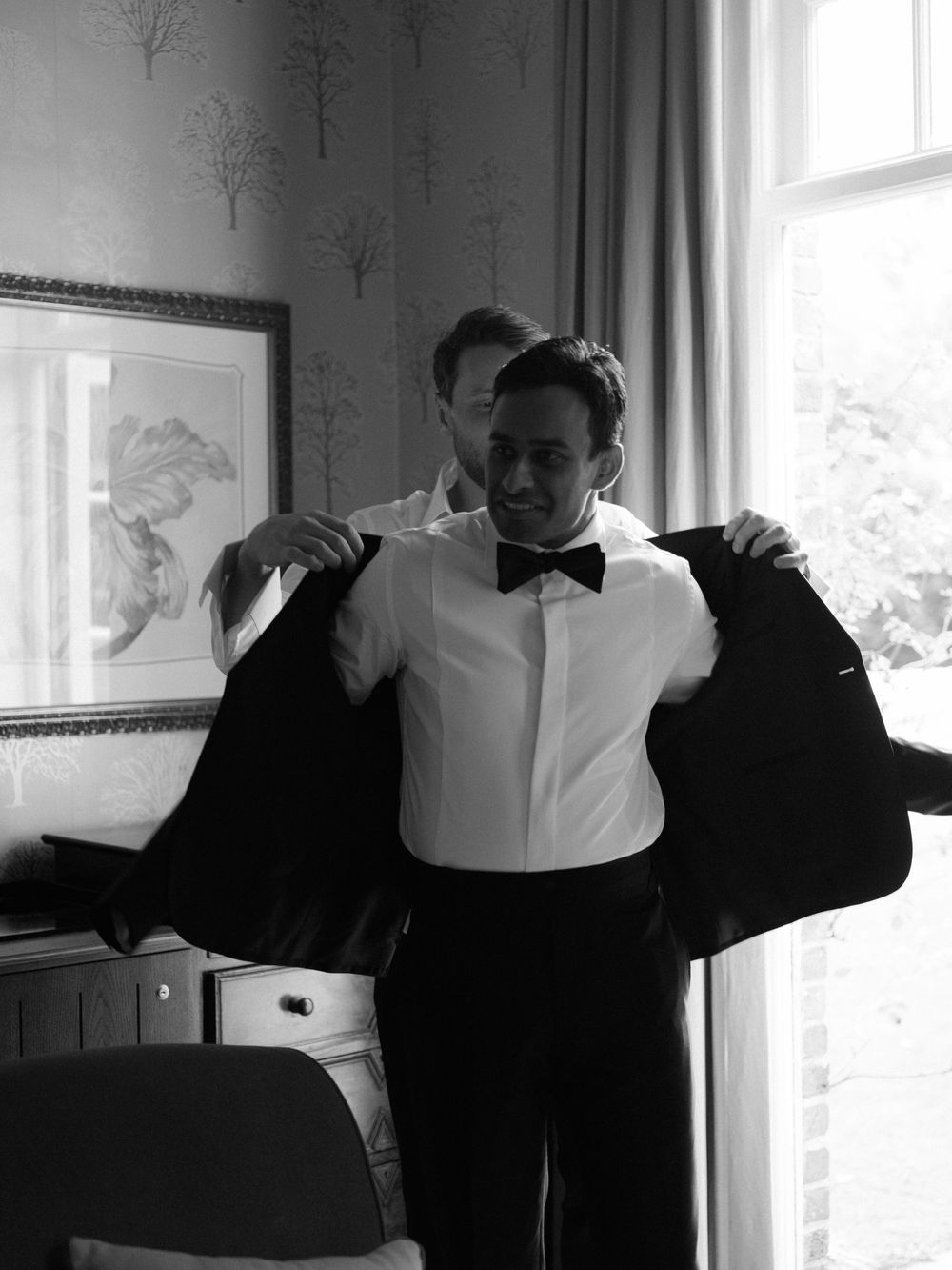 Groomsman helps groom put on black tuxedo jacket while getting ready indoors in black and white photo.